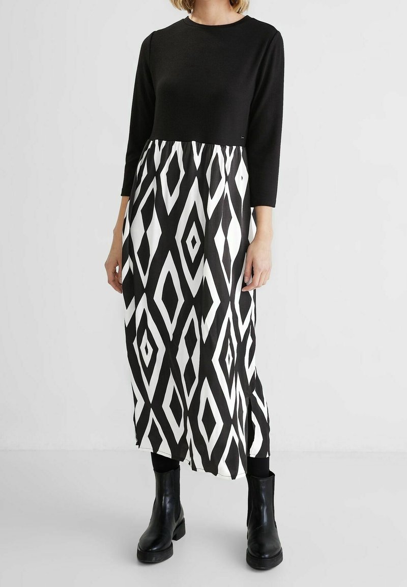 Femme portant une robe noire à manches longues avec une jupe à motif géométrique noir et blanc, des collants noirs et des bottines noires.