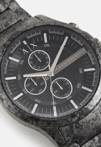 Montre analogique noire avec un cadran texturé, des accents argentés et trois sous-cadrans. Le bracelet a une finition noire à motif, lui conférant un aspect robuste.
