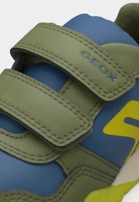 Μπλε και πράσινο αθλητικό παπούτσι με δύο πράσινους ιμάντες βέλκρο, το λογότυπο "GEOX" στον πάνω ιμάντα και μια κίτρινη λεπτομέρεια στο πλάι.