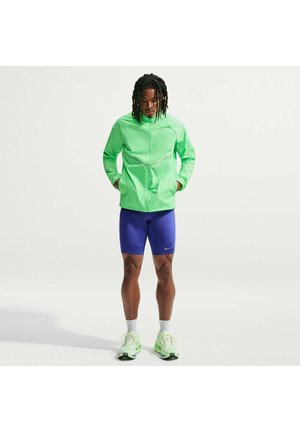 Homme portant une veste vert vif, un short de sport bleu et des chaussures de course vert clair, debout, regardant vers le bas les mains dans les poches.