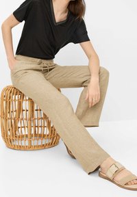 Czarna koszulka z dekoltem w serek z wąskim srebrnym wykończeniem, beżowe lniane spodnie z troczkiem oraz brązowe sandały z dużą klamrą.