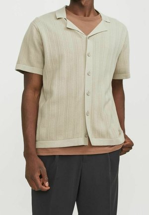 Camicia a maniche corte di un tenue beige con un motivo testurizzato, colletto morbido e chiusura frontale a bottoni. Indossata sopra una sottosovrapposizione marrone.