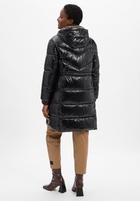 Schwarze, glänzende Daunenjacke mit Kapuze, die ein Chevron-Muster aufweist. Kombiniert mit braunen, engen Hosen und braunen Stiefeletten.