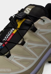 Bež sportska cipela s crnim detaljima, heksagonalnom teksturom i sistemom brzog vezivanja. Sadrži Gore-Tex oznaku i reflektirajuće dizajnerske elemente.