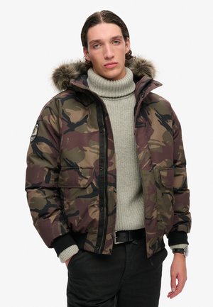 Chaqueta de plumas de camuflaje en tonos tierra, con capucha forrada de piel, cierre con cremallera y dos bolsillos delanteros, sobre un suéter de punto gris claro.