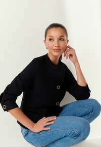 Čierna textúrovaná bunda so zlatými akcentmi a dvojitými gombíkmi, nosená cez modré denimové džínsy. Modelka je usadená na neutrálnom pozadí.