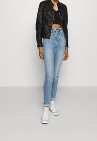 Persona vistiendo una chaqueta corta negra, un top corto negro, jeans ajustados de tiro alto en azul claro y zapatillas blancas, de pie sobre un fondo neutro.