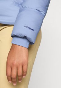 Hånd hvilende ved siden iført lyseblå Patagonia dunjakke med manchetten og beige bukser mod en ensartet baggrund.