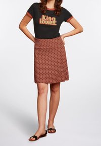 Woman wearing black graphic t-shirt with "King Louie" text, patterned knee-length skirt, black sandals, standing with hands on hips.