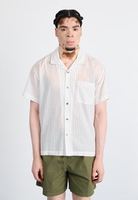 Kortermet, skjorte med knapper i gjennomsiktig hvitt stoff med smale vertikale striper og brystlomme, kombinert med olivengrønne shorts.