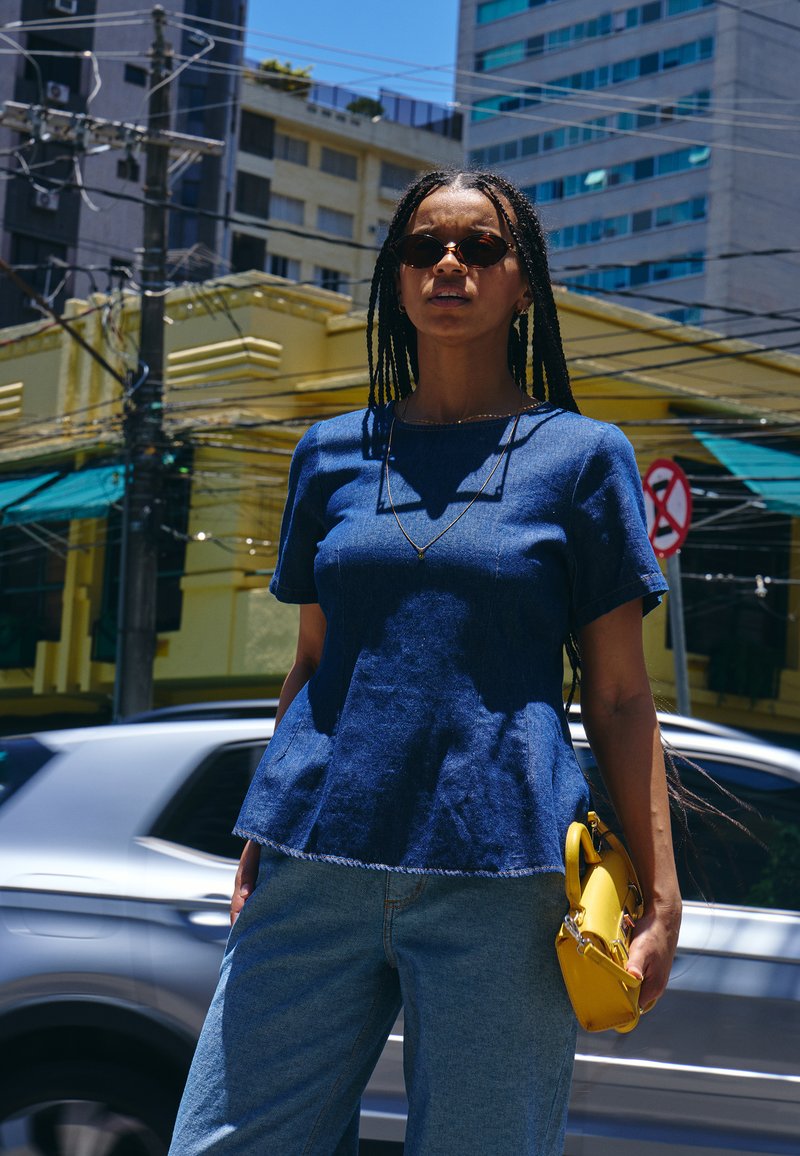 Femme aux longues nattes portant des lunettes de soleil, une chemise bleue et un jean, tenant un sac à main jaune, debout dans une rue de la ville avec des bâtiments et des voitures à proximité.