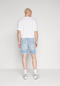 Pantalones cortos de denim azul claro con dobladillos enrollados, combinados con una camiseta blanca de corte simple. Costuras visibles y dos bolsillos traseros. Calzado casual completa el look.