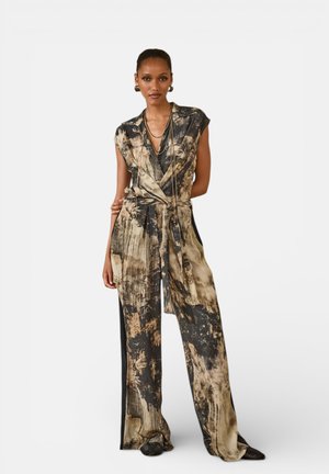 Donna che indossa una tuta senza maniche con motivo tie-dye, gambe larghe e cintura in vita, accessoriata con collane d'oro a strati e orecchini a cerchio.