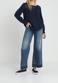 Marineblaues Langarmshirt mit strukturiertem Schultermotiv, kombiniert mit weiten blauen Jeans mit rohem Saum und schwarzen Sneakers.