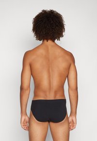 Schwarze Schwimmshorts aus dehnbarem Stoff, mit nahtlosem Design und glatter Textur, von hinten auf einem nackten Oberkörper gezeigt.