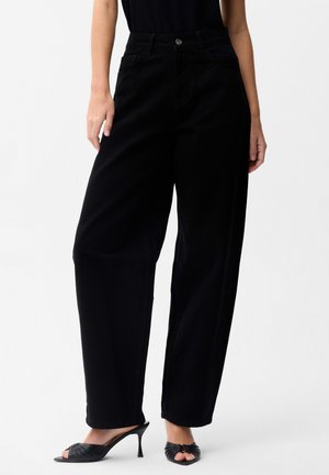 Femme portant un pantalon noir ample taille haute avec poches avant et des mules noires à bout ouvert à talons, debout devant un fond blanc.