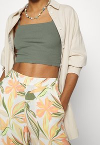 Haut court côtelé vert olive, chemise boutonnée beige surdimensionnée et pantalon large à motif floral dans des couleurs pastel avec des accents feuillus.