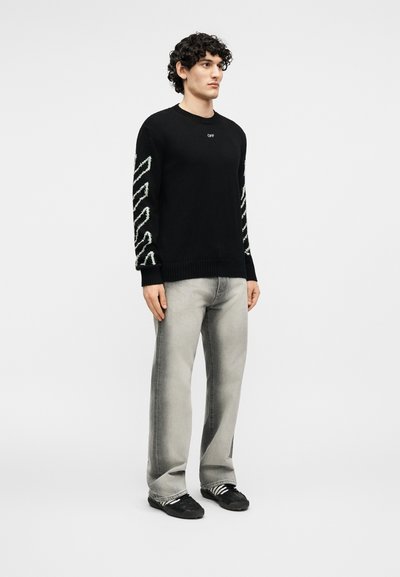 Νέος άνδρας που στέκεται φορώντας μαύρο πουλόβερ με λευκές διαγώνιες ρίγες στα μανίκια, ξεθωριασμένο γκρι τζιν και μαύρα sneakers με λευκές ρίγες.
