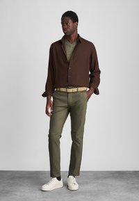 Camicia marrone con bottoni sopra una maglia verde, abbinata a pantaloni oliva e una cintura beige. Sneakers bianche completano il look. Sfondo semplice.
