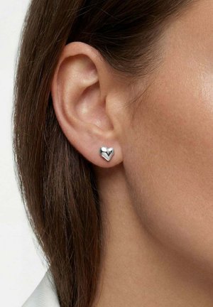 Närbild av en persons öra med rakt brunt hår, som bär ett litet glansigt silverörhänge i form av ett hjärta.