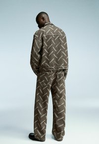 Jachetă și pantaloni cu model maro, confecționați dintr-un material texturat, având un design unic repetitiv și o croială lejeră. Pantofi negri completează ținuta.