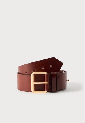 Une large ceinture en cuir brun rougeâtre est enroulée sur un fond blanc. Elle est dotée d’une grande boucle rectangulaire de couleur or et de plusieurs trous de réglage.