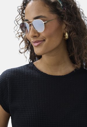 Frau mit lockigem Haar, die reflektierende Piloten-Sonnenbrillen, goldene Creolen und ein strukturiertes schwarzes Kurzarmoberteil trägt, leicht lächelnd.