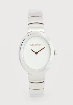 Dámske strieborné kovové náramkové hodinky Calvin Klein s oválnym ciferníkom, bielym ciferníkom, minimalistickým dizajnom a čiernymi hodinovými a minútovými ručičkami.