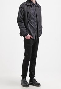 Giacca nera in nylon con colletto, chiusura a bottoni, sopra una camicia a quadri grigia. I pantaloni neri e le scarpe nere con i lacci completano l'outfit.