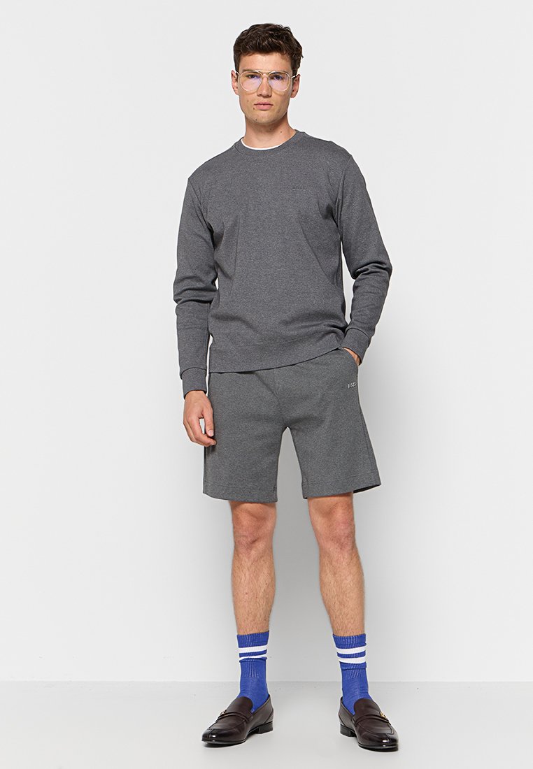 Sudadera gris de manga larga y cuello redondo, combinada con pantalones cortos grises a juego. Usada con calcetines azules con rayas blancas y zapatos oscuros.