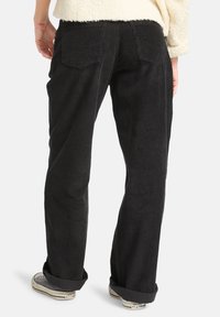 Pantalons en velours côtelé noir avec une coupe décontractée, présentant une texture côtelée verticale, deux poches arrière, et des ourlets retroussés sur des baskets blanches.