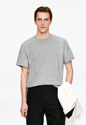 T-shirt gris en coton à manches courtes, col rond, coupe décontractée, associé à un pantalon noir et une veste légère posée sur un bras.