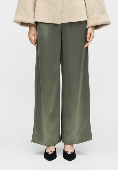 Μπεζ κοντό σακάκι πάνω από φαρδιά σατέν παντελόνια ελιάς με μαύρες μυτερές γόβες σε λευκό φόντο.