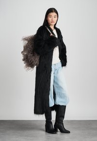 Cappotto lungo di pelliccia nera, camicia bianca, jeans a gamba larga di un blu chiaro e stivali neri in pelle fino al ginocchio. Accentuato da una borsa trasparente stampata.