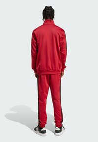 Sarkans sporta uzvalks ar melniem sānu svītrām. Izgatavots no gludas, spīdīgas materiāla. Iekļauj augstu apkakli un pielāgotas aproces. Melnas sporta apavi.