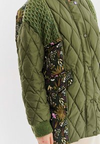 Prošívaná olivově zelená bunda s pletenými záplatami na ramenou a květinovým vzorem na rukávech a bocích.