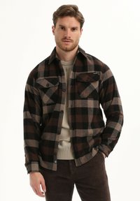 Flanellen shirt in zwart, bruin en beige geruit patroon; beschikt over twee borstzakken, knoopsluiting en kraag. Draagt over een lichte trui.