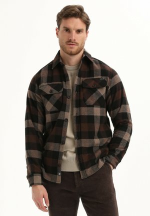 Flanellen shirt in zwart, bruin en beige geruit patroon; beschikt over twee borstzakken, knoopsluiting en kraag. Draagt over een lichte trui.
