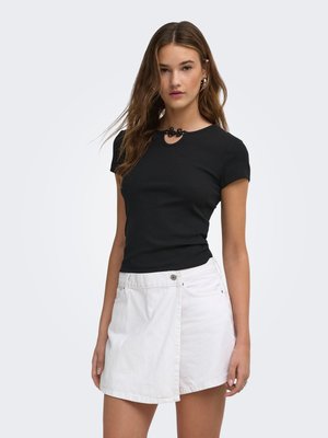 Jeune femme aux longs cheveux bruns portant un haut noir à manches courtes avec un col décoratif et une mini-jupe asymétrique blanche sur un fond uni.