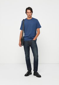 Blue short-sleeve t-shirt, round neck, paired with dark denim jeans and black shoes. Model stands with a black crossbody bag.