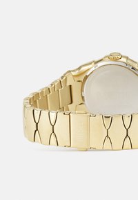 Montre en acier inoxydable doré avec un design de bracelet chunky à maillons. Elle présente un boîtier rond, un verre clair et une couronne visible sur le côté.