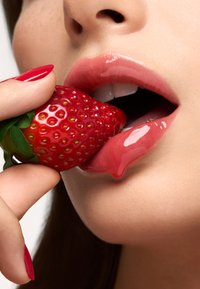 Close-up van een glanzende, roze lip met een verse, rode aardbei die wordt vastgehouden door een verzorgde hand met rode nagels, waarbij de sappige textuur en glans worden getoond.