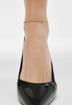 Zapato de tacón alto de charol negro con punta afilada, con un delicado tobillo dorado alrededor del tobillo. Textura suave, diseño minimalista.