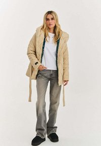 Béžová prošívaná bunda s kapucí, s zelenou vzorovanou podšívkou. Kombinována s bílou košilí, šedými rozšířenými džínami a černými nazouvacími botami.