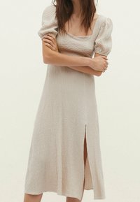 Cremfarbendes Kleid mit quadratischem Ausschnitt, Puffärmeln, strukturiertem Stoff und einem seitlichen Schlitz für zusätzliche Details. Knielanger Schnitt.