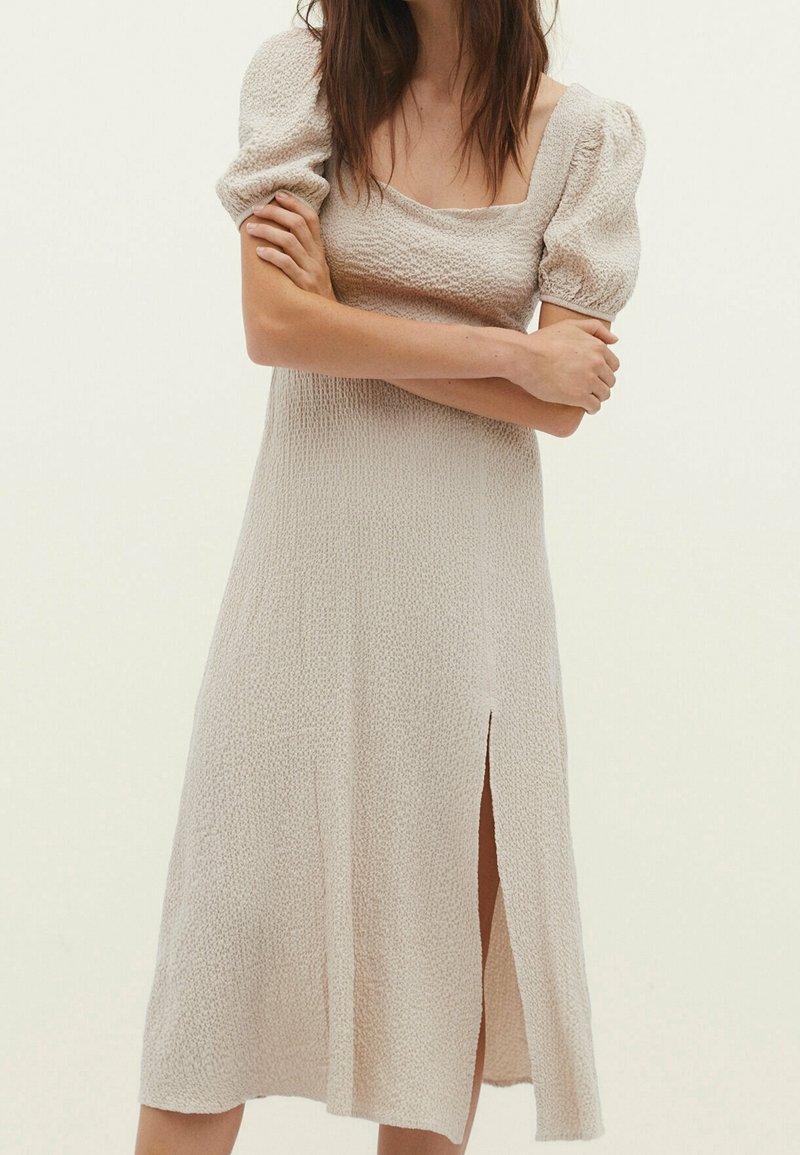 Cremfarbendes Kleid mit quadratischem Ausschnitt, Puffärmeln, strukturiertem Stoff und einem seitlichen Schlitz für zusätzliche Details. Knielanger Schnitt.