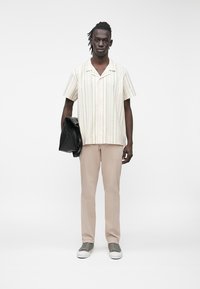 Trumpomis rankovėmis, kreminės spalvos marškiniai su vertikaliomis juostomis, derinami su smėlio spalvos kelnėmis ir pilkais sportbačiais, laikantys juodą švarką.