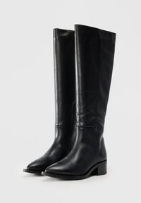Bottes en cuir noir hautes avec de petits talons carrés et des fermetures éclairs latérales, présentées debout sur un fond neutre.