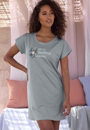 Woman in gray dress with short sleeves stands indoors near cushions, dress front text reads "let your dreams bloom" with flower graphic.