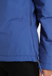 Detailní záběr osoby v modré bundě se zipem na boku a černých kalhotách, zobrazující detaily švů na rukávu a spodním lemu.